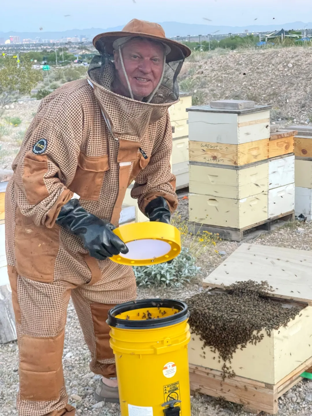 A beekeeper in a brown suit holding a yellow lid over a bucket, with hives behind