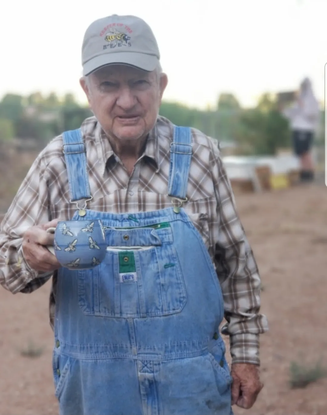 An older beekeeper in denim overalls and a bee cap, holding a mug painted with bees